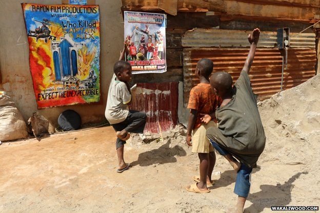 Children do kung fu poses in front of posters advertising the next Wakaliwood film