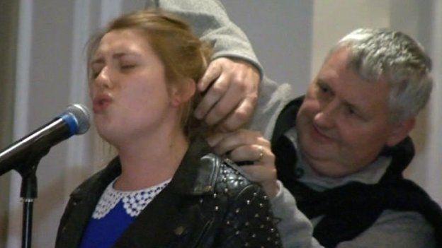 Ella Henderson singing into a mic while her father Sean ties her hair