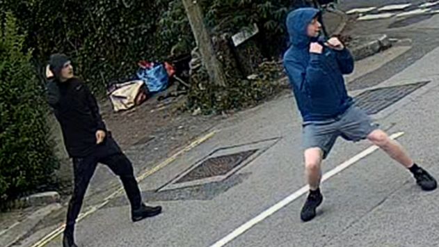 Police CCTV image of two men - one wearing all-black clothing, including a hooded jacket; the other wearing light blue shorts, black trainers and a dark blue coat. The latter is stood in a fighting stance with clenched fists, looking towards something out of shot.