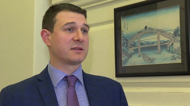 Matt Binley with short dark hair wearing a purple tie and blue jacket next to  a wall with a picture of a bridge on display