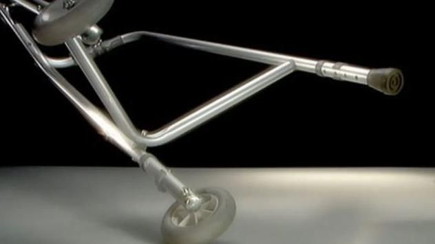 A silver-coloured walking aid with wheels held at an angle against a black background.