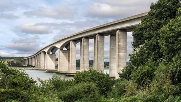 The Orwell Bridge in Suffolk, an image taken from a bank by the side of the River Orwell. The bridge is large, grey structure with a river running beneath it, with green foliage by it.