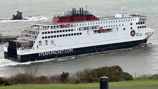 The Manxman ferry leaving Douglas Harbour. The ferry is painted red, white and black and has STEAM-PACKET.COM written on the side of the hull. The green grass of Douglas Head can be seen in the fore,  while the castle-like structure of the Tower of Refuge can be seen behind the ferry in the bay.