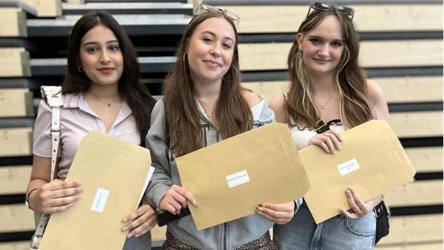 Students collecting their A Level results at All Saints Academy in Cheltenham. Three young woman stand alongside each other, all of them holding a brown A4 envelope