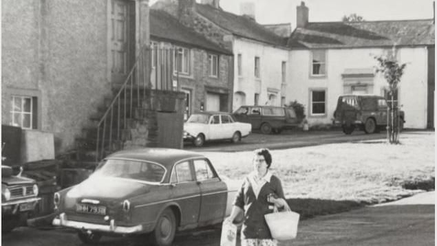 Black and white picture of woman in the 1960s walking through the village in front of the shop while carrying shopping bags