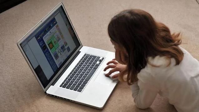 A young girl lying on a carpeted floor using a silver laptop