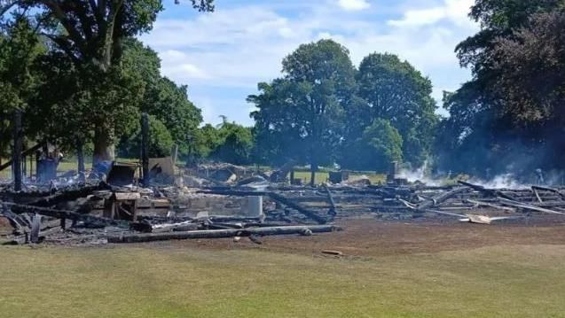 On a large patch of grass, the clubhouse site is gone, replaced with burning timber and the embers of the former structure, with smoke rising from the beams. 