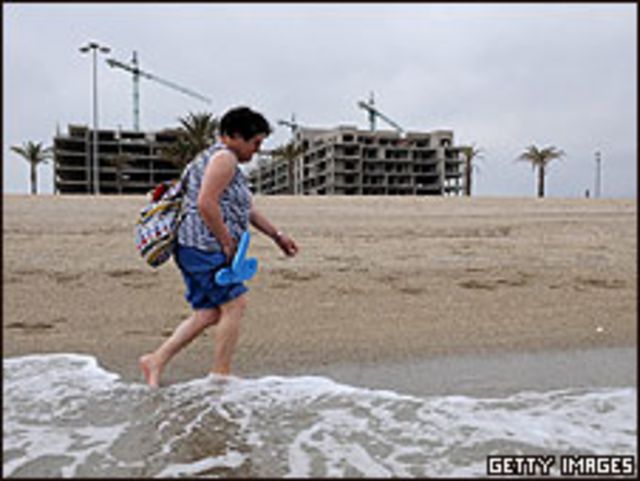 Una mujer camina por la playa; construcciones abandonadas detrás