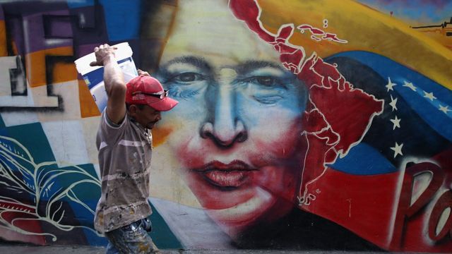 Hombre caminando en Caracas frente a un mural de Hugo Chávez.
