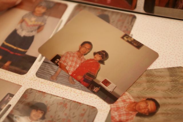 Fotografia colorida mostra um album de fotos com fotos de família, em primeiro plano há uma menina pequena no colo de um homem de camisa xadrez