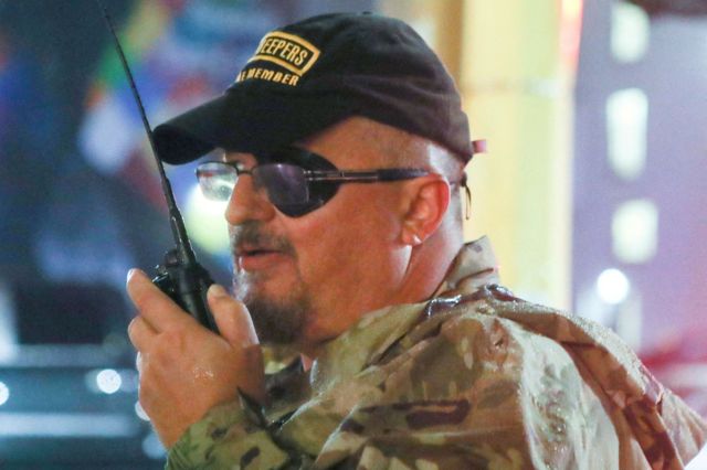 Stewart Rhodes, um homem branco de meia idade, de bigode e boné, usando um tapa olho e um walkie talkie