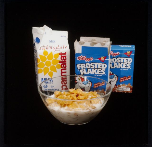 A bowl of cereal with milk, a milk carton and two packets of Frosted Flakes