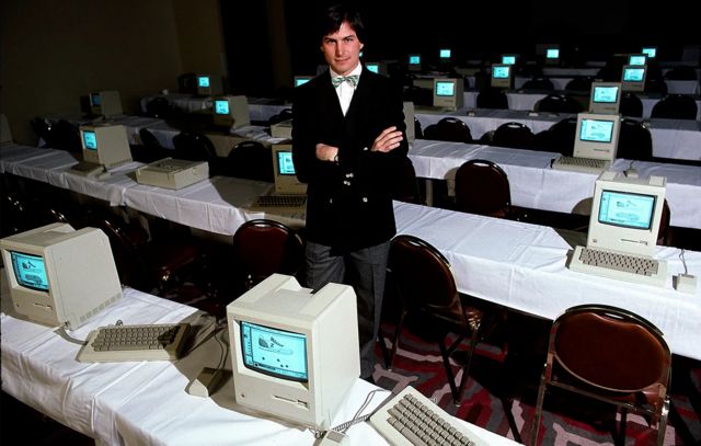 Steve Jobs de braços cruzados com olhar confiante entre mesas com computadores Macintosh