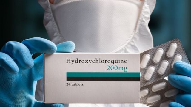 Profesional de la salud con mascarilla sosteniendo una caja de "Hydroxychloroquine 200 mg y un blíster con píldoras blancas.