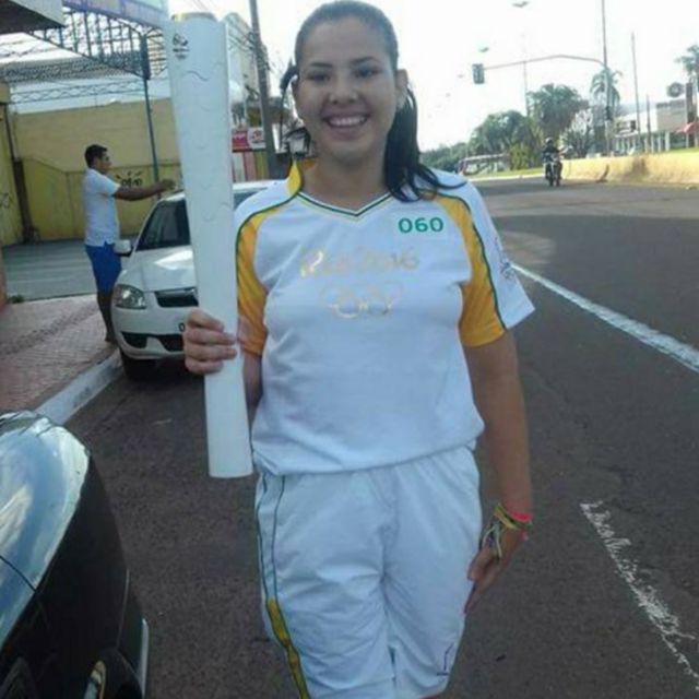 A então estudante Camila Micaela de Oliveira Fonseca segura com a mão direita uma tocha olímpica ainda apagada. Ela veste uniforme branco com bermuda e calça tênis vermelhos