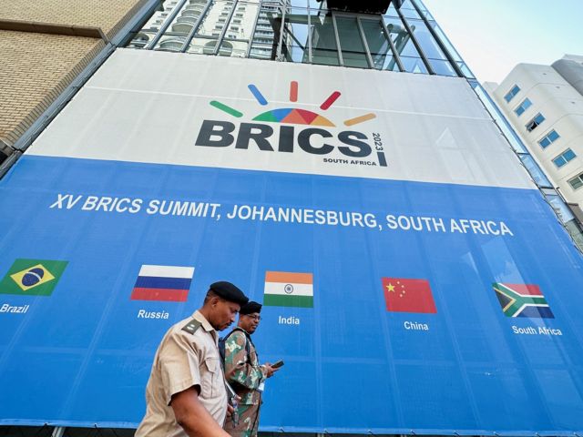 Dois homens passam em frente a painel de evento dos BRICS que mostra cinco bandeiras dos membros