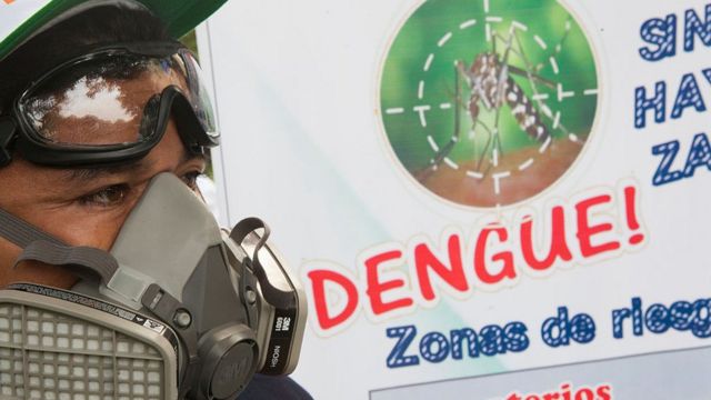 Un hombre con una máscara protectora pasa frente a un cartel que advierte del dengue