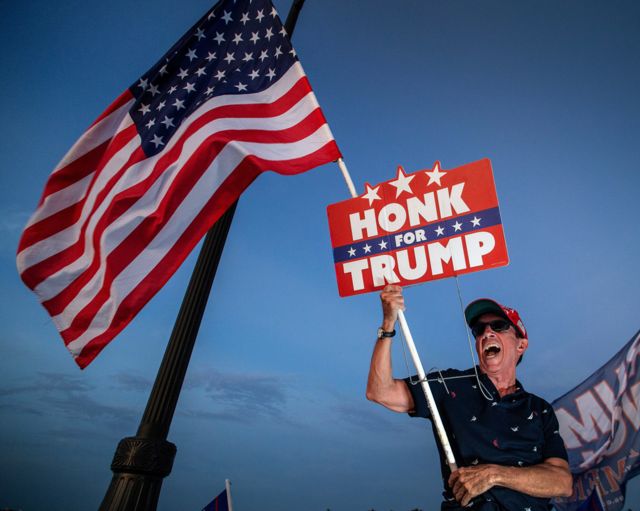 Fotografia colorida mostra um homem branco de meia idade segurando uma bandeira americana e um placa onde está escrito "buzine por Trump".