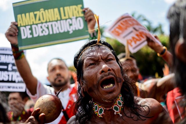 Indígena gritando em protesto, com cartazes atrás pedindo proteção à Amazônia