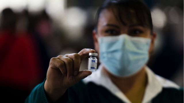 Mulher com traços hispânicos e usando máscara aparece desfocada enquanto exibe frasco de vacina à frente do rosto
