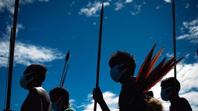 Indígenas Yanomami enfileirados na contraluz, com céu azul atrás