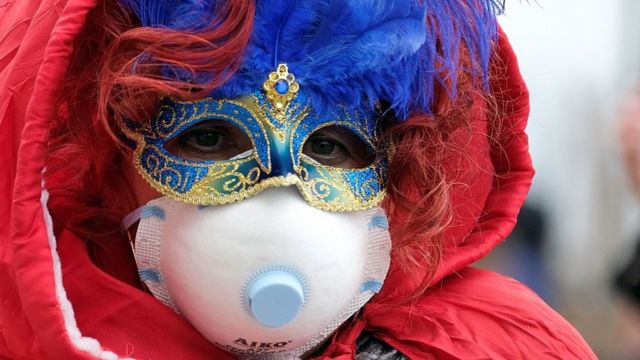 Un asistente al carnaval de Venecia usando una mascarilla protectora junto a su disfraz.