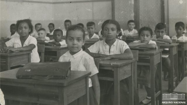 Crianças em escola no Rio, sentadas em suas cadeiras, olham para câmera 