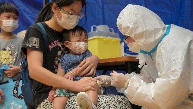 A medical worker wearing a full set of personal protective equipment vaccinates a young child at a vaccination center in Hong Kong (Photo by China News Agency 28/8/2022)