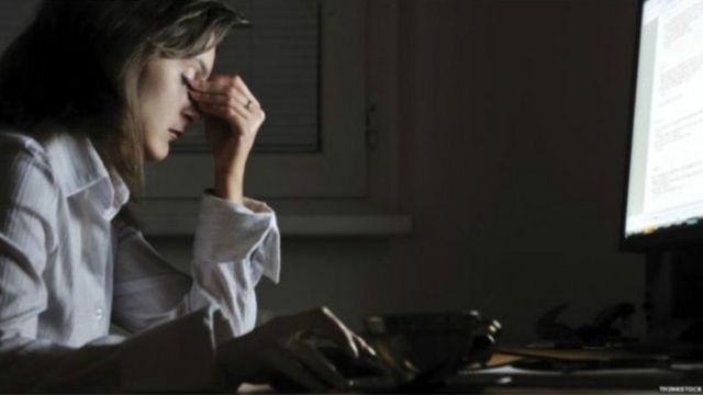 Mulher em frente ao computador parece estressada, com os olhos fechados e os dedos sobre os olhos
