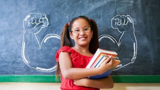 Menina de pele parda sorridente segurando livros em frente a braços desenhados na lousa