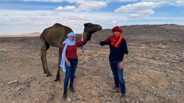 Casal posa para foto ao lado de camelo em terreno árido