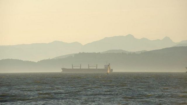Um veleiro passa junto a um navio de carga em English Bay com as montanhas da costa ao fundo, perto do centro de Vancouver