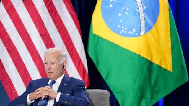 Biden sentado com olhar sério e dedos cruzados em frente a bandeiras dos EUA e Brasil