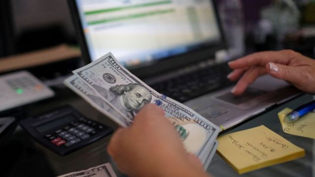 Una mujer sostiene unos dólares en su mano izquierda mientras trabaja en su computador.