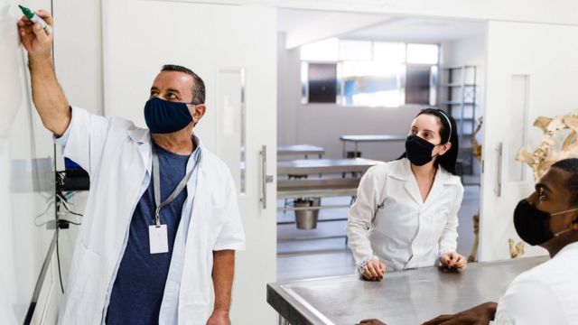 Instrutor escreve em quadro branco, observado por dois alunos sentados diante de mesa, em ambiente de laboratório