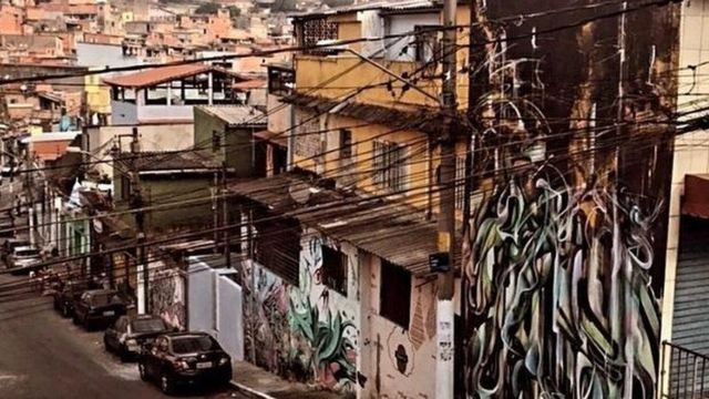 Imagem mostra uma rua de Vila Flavia, comunidade do distrito de São Mateus, na Zona Leste de São Paulo. Há uma fileira de carros estacionados, casas com grafites nos muros e, ao fundo, casas de tijolo aparente, típicas das periferias