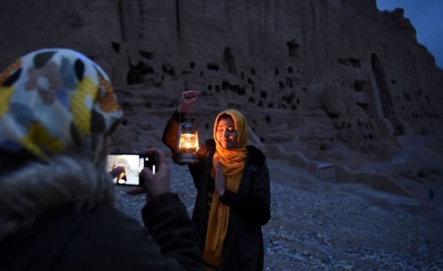 An Afghan woman holding a lantern poses in the dusk against a carved mountainside