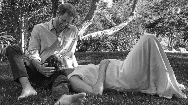 Em foto em preto e branco, Meghan deita no colo de Harry, que está sentado embaixo de uma árvore