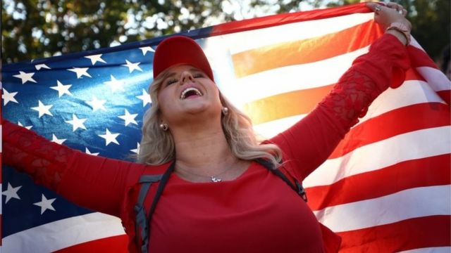 Mulher sorri de olhos fechados enquanto ergue bandeira americana por trás da cabeça em protesto em Washington