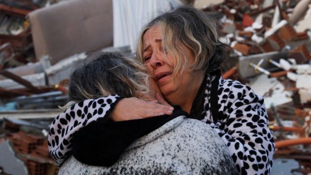 Mulheres se abraçando e chorando com escombros ao fundo