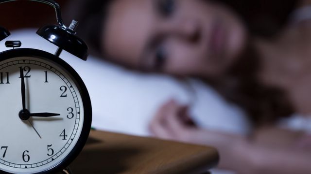 Close up of an alarm clock with a woman lying in bed in the background.