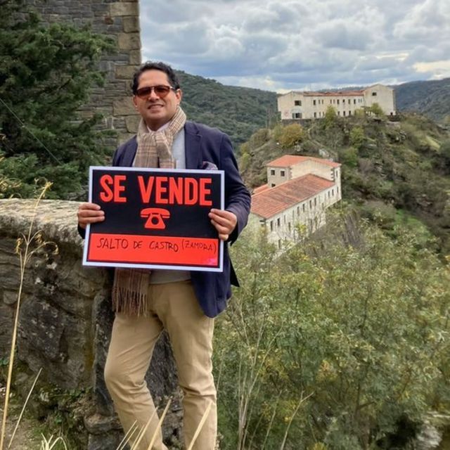 Ronnie Rodriguez with a sign of "For Sale" and the village in the background