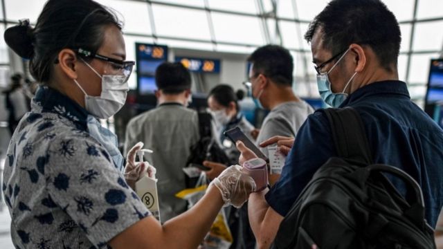 Funcionária de aeroporto em Wuhan checa temperatura de passageiro, ambos de máscaras