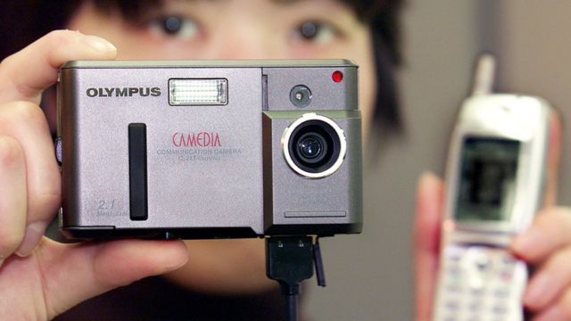 An out-of-focus person holds an Olympus Camedia camera in one hand, an early blocky mobile phone in another, with a cable running between them