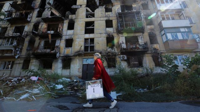 在乌克兰马里乌波尔，一名妇女提着便携选票袋经过一座严重受损的居民楼。(photo:BBC)