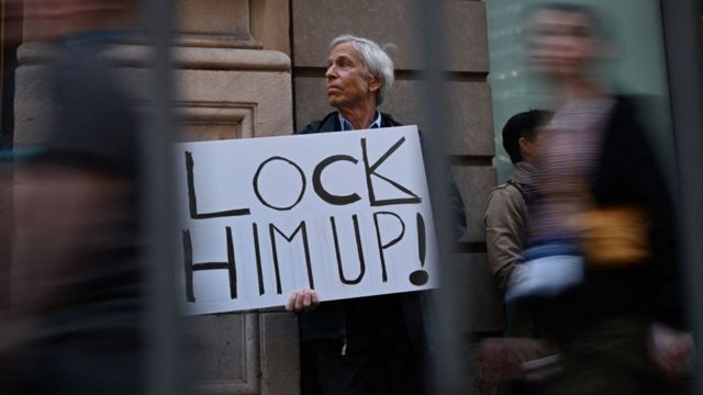 A protester holds a sign reading "Lock Him Up!" as he stands outside of a media area near Trump Tower