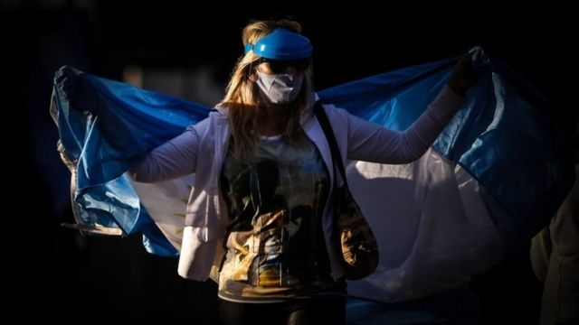 Mulher protesta usando máscara e com uma bandeira da Argentina em volta do corpo
