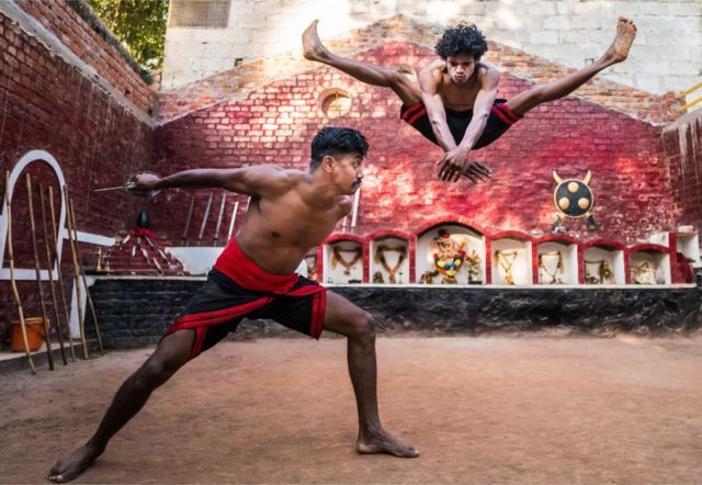 Dos hombres realizan una secuencia de artes marciales, uno enarbola un cuchillo mientras el otro da un gran salto en el aire para esquivarlo