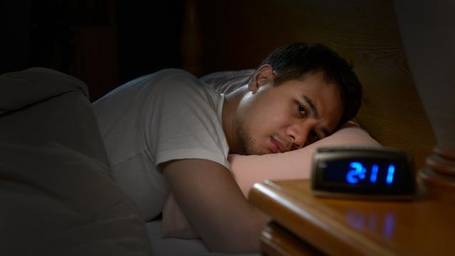 Jovem latino deitado na cama olha para o relógio, que mostra que é madrugada, com olhar de preocupação