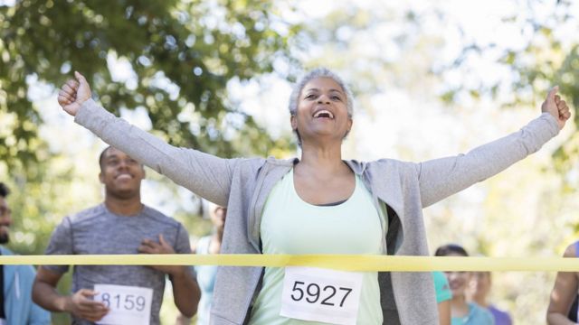 Mulher sorri e ergue os braços ao enconstar na linha de chegada de uma maratona